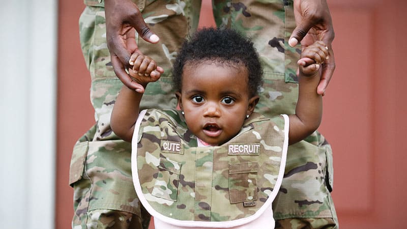 baby learning to walk is held by hands by her mom in military uniform