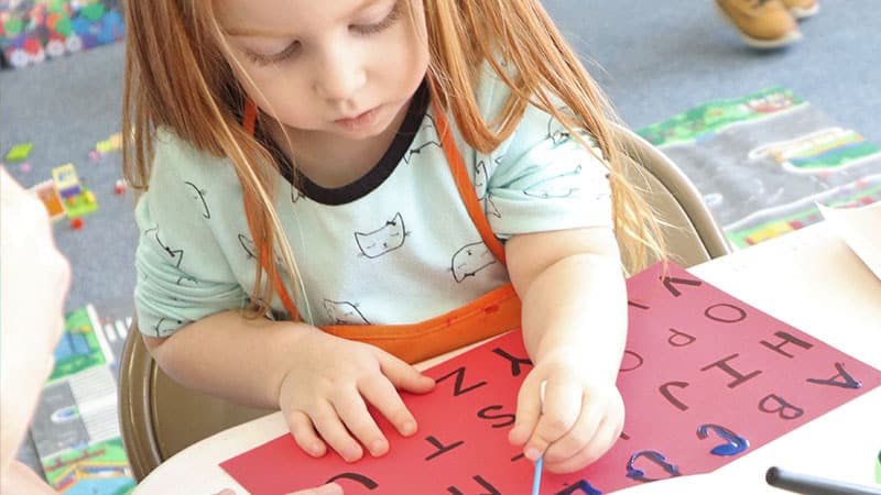a little girl practices her letters with her mom’s hand holding the paper down